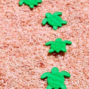 May include: Four small, bright green turtle-shaped figures are arranged in a diagonal line across a bed of light pink, loose grains. Each turtle figure features a detailed shell pattern with a head, four flippers, and a tail clearly visible.