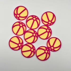 May include: A collection of ten yellow and magenta beach ball-shaped embellishments. Each circular piece features a yellow center with magenta lines, mimicking a beach ball design. The embellishments are arranged on a white surface.