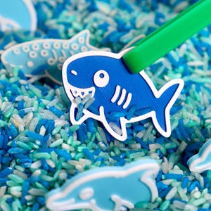 May include: A bright blue shark-shaped figure with white outlines being held by green plastic tweezers, positioned over a bed of loose, multi-colored blue and white rice grains, with other light blue sea creature figures scattered throughout.