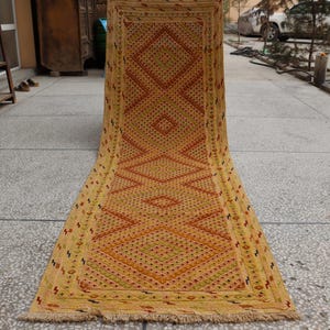 May include: A rectangular, golden-yellow rug with a geometric pattern of diamonds and zigzags in shades of red, orange, and green. The rug has a fringed edge and is displayed outdoors on a concrete surface.