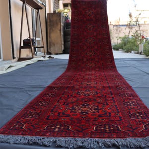 May include: A long, narrow runner rug featuring a deep red background with intricate geometric patterns in black, navy blue, and cream. The rug has a repeating medallion design throughout its length and is finished with short, light grey fringe at the ends.