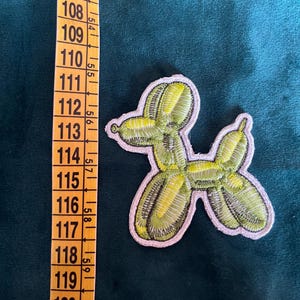 Può includere: Toppa ricamata a forma di cane palloncino, con sfumature di verde e giallo e un bordo bianco. La toppa è posizionata su un tessuto verde scuro accanto a un metro a nastro.