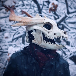 May include: A detailed, off-white skull mask with prominent teeth and large eye sockets. The mask has attached, curved, tan-colored horns. The person is wearing a black jacket and a red scarf, set against a snowy, blurred background.