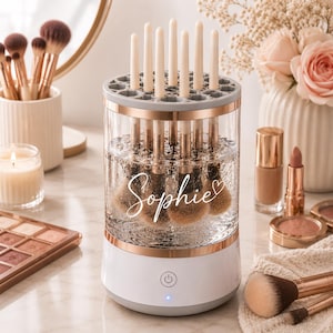 Op de afbeelding: Een cilindrische make-up kwastenreiniger met een witte basis, een doorzichtig middengedeelte en een grijze bovenkant met meerdere sleuven. Het doorzichtige gedeelte toont de naam Sophie in wit handschrift met een hartvormig icoon.