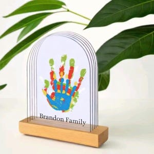 Puede incluir: Una obra de arte con huella de mano, con base de madera y marco arqueado transparente. La huella de mano está hecha con pintura colorida, incluyendo azul, verde, rojo y amarillo. El texto "Brandon Family" está impreso debajo.