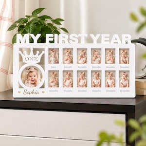 Può includere: Una cornice portafoto rettangolare in legno bianco con la scritta MY FIRST YEAR nella parte superiore. Include un'ampia apertura circolare sormontata da un intaglio a forma di corona e dodici piccoli scomparti rettangolari etichettati da born a 11months. Il nome Sophia è stampato in corsivo con due piccoli cuori.