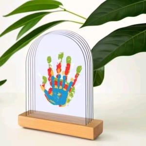 May include: A colourful handprint artwork displayed in a clear, arched frame with a wooden base. The handprint is made with red, green, blue, and yellow paint. The frame is supported by thin, vertical metal rods.