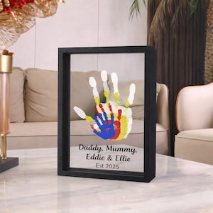 May include: A framed handprint artwork in a black frame. The handprint is created with white, yellow, red, and blue paint. The text "Daddy, Mummy, Eddie & Ellie Est 2025" is printed below the handprint.