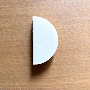 Può includere: Una forma di formaggio a mezzaluna, bianco sporco, con una superficie leggermente ruvida. Il formaggio è appoggiato su uno sfondo di legno marrone chiaro, creando un contrasto di colore e consistenza. Il formaggio sembra fresco e pronto da mangiare.
