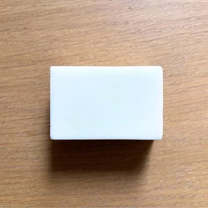 May include: A rectangular, white eraser is centered on a wooden surface. The eraser is a solid, opaque white and has a smooth, clean appearance. The wood grain of the surface is visible, with warm brown tones.