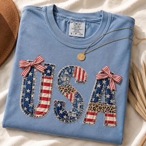 May include: Light blue t-shirt with the word "USA" appliqued in a patchwork design featuring the American flag and leopard print. Red and white striped bows are attached to the letters. A gold necklace and a brown hat are also visible.