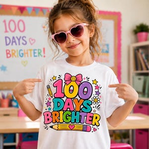 May include: A girl wearing a white t-shirt with the text "100 DAYS BRIGHTER" in bright colours. The shirt design includes a pencil, stars, and a bow. She is wearing heart-shaped sunglasses and pointing at the shirt.