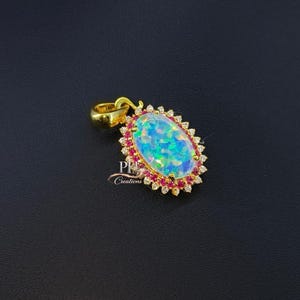May include: A gold pendant with an oval, iridescent opal center stone. The opal is surrounded by a row of small red gemstones and a border of small, clear gemstones. The pendant has a gold bail.