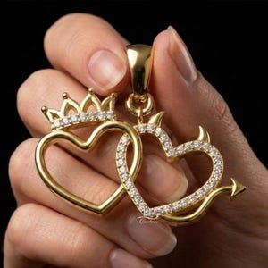 May include: A gold pendant featuring two interlocking heart shapes. The left heart is topped with a small crown, while the right heart features devil horns and a pointed tail. Both hearts are outlined with small, clear, sparkling stones.