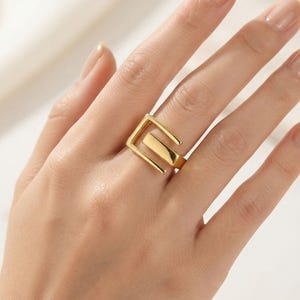 May include: A gold-toned ring featuring an open, geometric design with a central rectangular bar and two parallel angular arms that extend to form a square-like shape. The metal has a smooth, polished finish that reflects light across its surface.