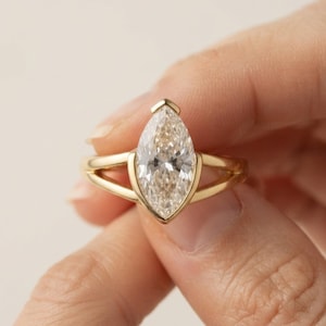 Puede incluir: Un anillo de oro con una piedra preciosa transparente de corte marquesa montada en un engaste en forma de V. La banda está dividida en dos segmentos dorados finos y paralelos que se unen en la base de la piedra central.