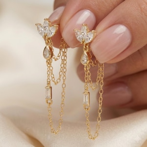 Puede incluir: Un par de pendientes dorados con un poste en forma de flor con piedras transparentes de corte marquesa, una piedra colgante en forma de lágrima y dos cadenas delicadas conectadas a una pequeña piedra rectangular de corte baguette.