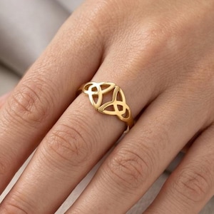 May include: A gold ring featuring a central openwork design composed of three interconnected loops forming a triquetra shape. The band is thin and polished, showcasing a metallic yellow finish that sits prominently on the finger.
