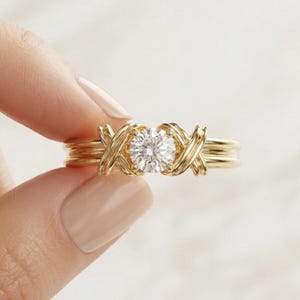 May include: A gold ring featuring a central round brilliant-cut clear gemstone. The band is composed of multiple thin, polished gold wires that cross over each other in an X-shaped pattern on both sides of the central stone setting.