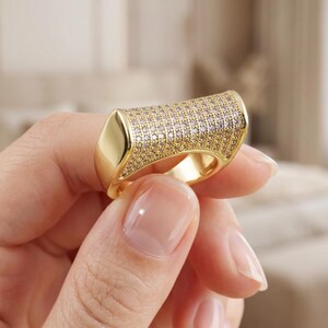 May include: A gold-toned ring featuring a wide, rectangular top surface densely encrusted with numerous small, clear, sparkling stones. The band has a smooth, polished finish that contrasts with the textured, stone-covered top section.
