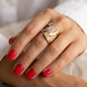 May include: A two-tone open ring featuring a silver-colored upper section and a gold-colored lower section, both designed with a series of parallel, wavy, grooved lines that create a textured, layered appearance across the finger.