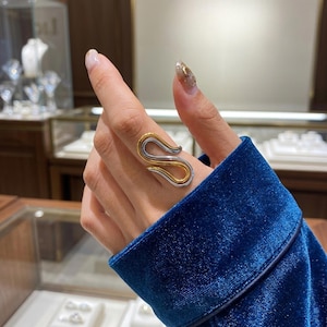 May include: A two-tone ring featuring a fluid, S-shaped design. The ring is constructed with a polished gold-colored inner band nested within a smooth, silver-colored outer band, creating a layered, continuous loop appearance on the finger.
