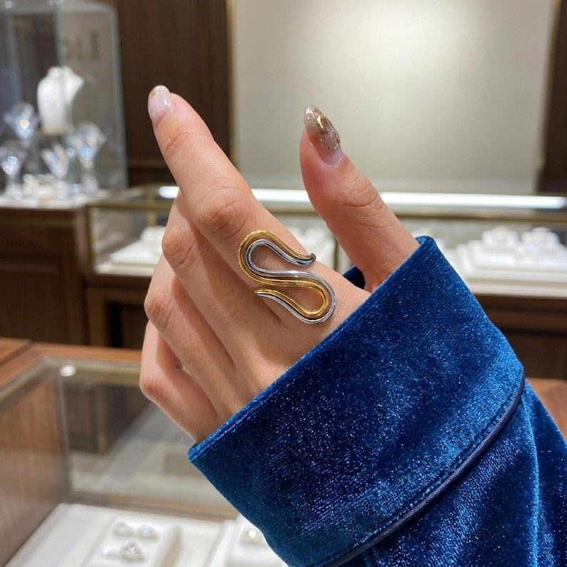 May include: A two-tone ring featuring a fluid, S-shaped design. The ring is constructed with a polished gold-colored inner band nested within a smooth, silver-colored outer band, creating a layered, continuous loop appearance on the finger.