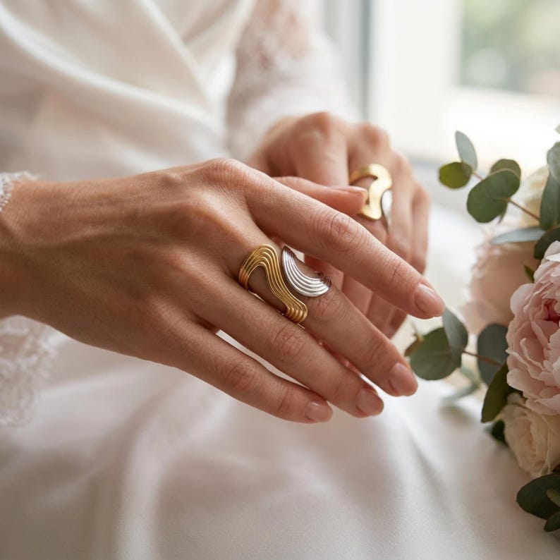 May include: A two-tone ring featuring a gold-colored section with parallel wavy lines and a silver-colored section with a textured, fan-like pattern. The ring is worn on a finger, and a second, similar gold-colored ring is visible on another finger.