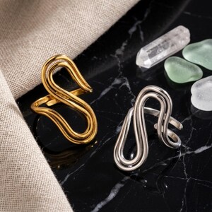May include: Two open-style rings featuring a continuous, fluid, S-shaped wire design. One ring is finished in a polished gold tone, while the other is finished in a polished silver tone. Both rings have an adjustable band with an open back.