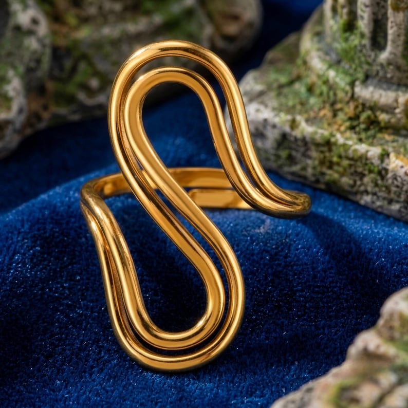 May include: A gold-toned ring featuring a continuous, fluid, open-loop design that curves into an elongated S-shape. The metal surface is smooth and polished, creating a reflective finish that highlights the interconnected, swirling wire-like structure.
