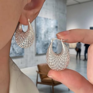 May include: A pair of silver hoop earrings featuring a hollow, teardrop-shaped design with an intricate, open-work grid pattern. The earrings have a polished metallic finish and a hinged post closure, showcasing a lightweight, cage-like structure.