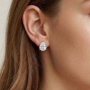 Może przedstawiać: Zbliżenie ucha z kolczykiem z diamentem w kształcie łzy. Przezroczysty diament błyszczy w złotej oprawie. Kolczyk to klasyczna i elegancka biżuteria.