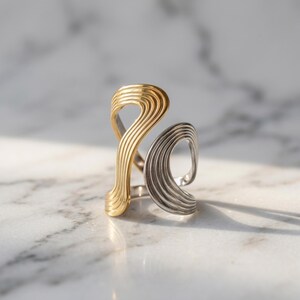 May include: A two-tone ring featuring a gold-colored section and a silver-colored section, both designed with a series of parallel, raised ridges that create an open, flowing, and asymmetrical shape that wraps around the finger.