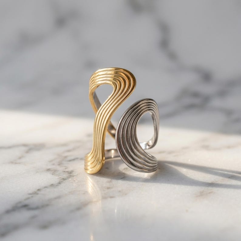 May include: A two-tone ring featuring a gold-colored section and a silver-colored section, both designed with a series of parallel, raised ridges that create an open, flowing, and asymmetrical shape that wraps around the finger.