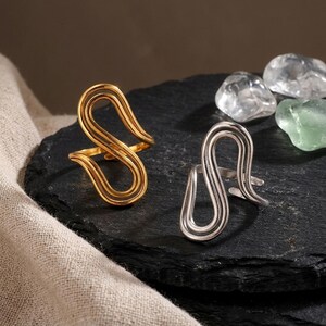 May include: Two open-style rings featuring a continuous, fluid, S-shaped wire design. One ring is finished in a polished gold tone, while the other is finished in a polished silver tone. Both rings have an adjustable band structure.