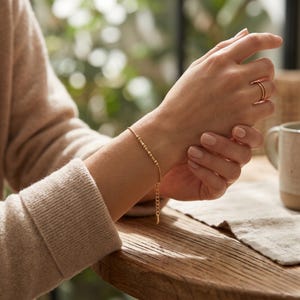 May include: A delicate gold bracelet with small, evenly spaced beads and a chain extension. The bracelet is worn on a wrist, with a matching gold ring on a finger. The background is softly blurred, suggesting a natural setting.