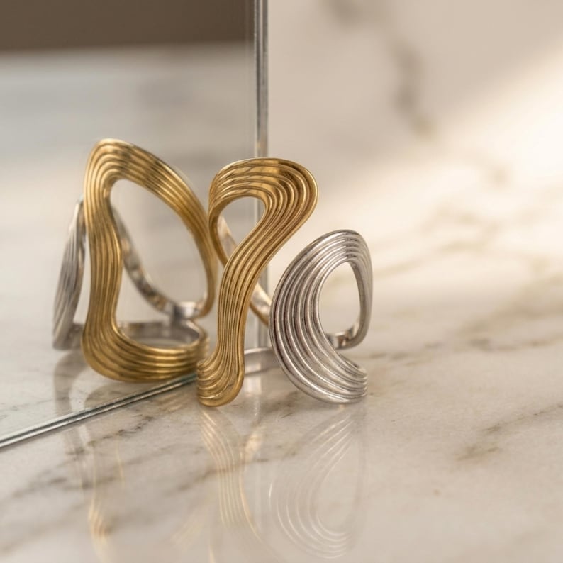 May include: A gold-toned ring and a silver-toned ring, both featuring an open, fluid, and wavy design with parallel grooved lines. The rings are positioned near a reflective surface, creating a mirrored visual effect of the metallic jewelry pieces.