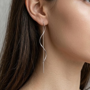 Peut inclure: Une longue boucle d'oreille en argent de type fil, dotée d'un design métallique ondulé et délicat qui pend verticalement depuis le lobe. La boucle se compose d'une fine chaîne en argent poli reliée à une tige métallique courbe et sinueuse.