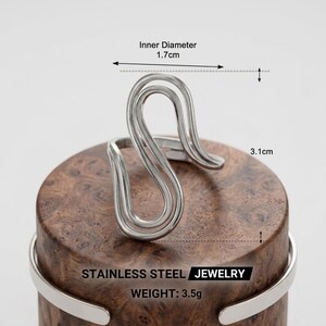 May include: A silver-toned stainless steel ring with a wavy, open-ended design. The ring features an inner diameter of 1.7 cm and a total height of 3.1 cm. The text on the image reads STAINLESS STEEL JEWELRY and WEIGHT: 3.5g.