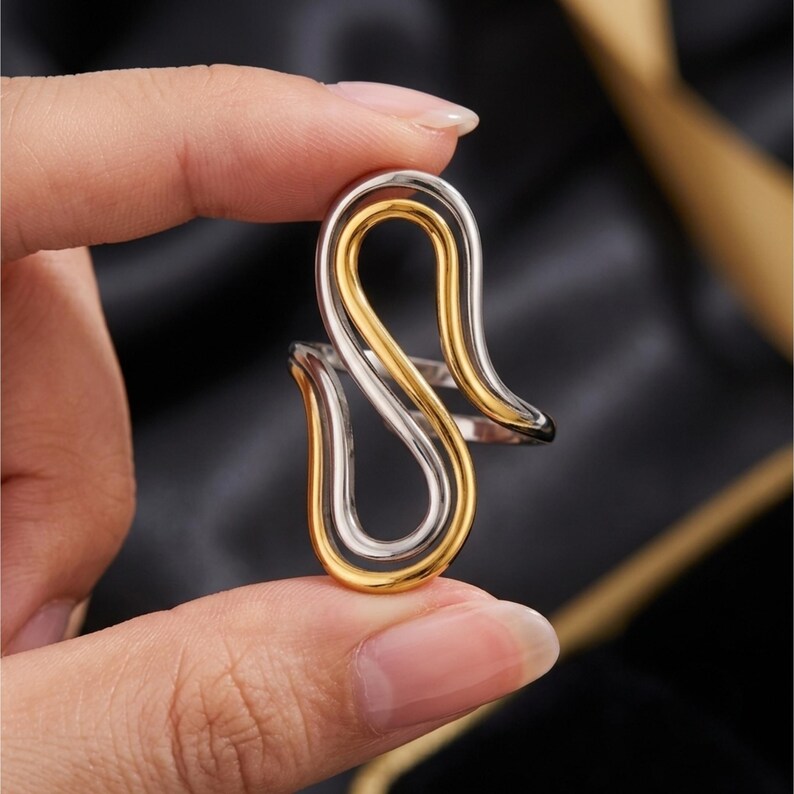 May include: A two-tone ring featuring a fluid, open-loop design with a polished silver-colored band intertwined with a smooth gold-colored band. The continuous, curved metal lines create an elongated, abstract shape that wraps around the finger.