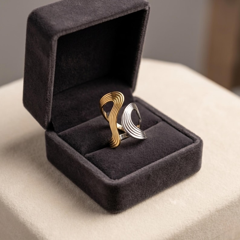 May include: A two-tone ring featuring a gold-colored section and a silver-colored section, both with a wavy, ribbed texture. The ring is displayed inside a dark gray, velvet-lined jewelry box with a hinged lid that is open.