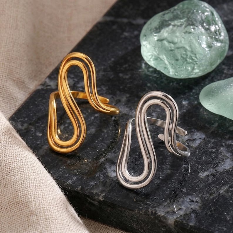 May include: Two open-style rings featuring a continuous, fluid, wave-like wire design. One ring is finished in a polished gold tone, while the other is finished in a polished silver tone. Both rings have an adjustable band structure.