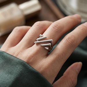 May include: A polished silver ring featuring a geometric design composed of three stacked, angular, chevron-shaped bars that create a layered, stepped appearance. The metal has a smooth, reflective finish and a clean, structural form.