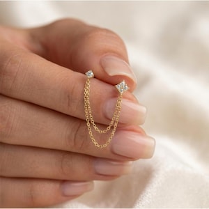 Puede incluir: Un pendiente de doble cadena dorada con dos gemas cuadradas y transparentes. El pendiente está sujeto por una mano con uñas cuidadas, sobre un fondo suave y borroso. El diseño incluye un pequeño perno y una cadena colgante.
