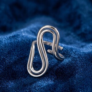 May include: A silver-toned ring featuring a continuous, fluid, double-loop design that creates an elongated, abstract shape. The metal band is polished and reflective, with an open-ended structure that allows for adjustment in size.