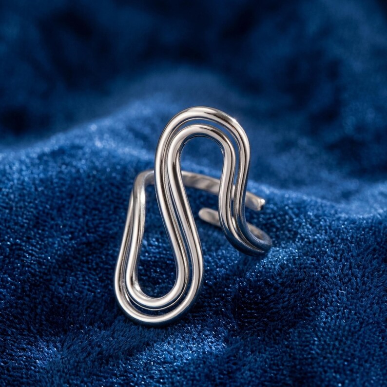May include: A silver-toned ring featuring a continuous, fluid, double-loop design that creates an elongated, abstract shape. The metal band is polished and reflective, with an open-ended structure that allows for adjustment in size.