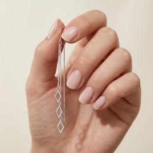 May include: A silver threader earring featuring a delicate chain design with three open diamond-shaped charms hanging vertically. The earring is constructed from polished silver-toned metal, showcasing a minimalist geometric aesthetic with a long, slender chain that loops through the ear.