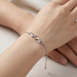 Könnte beinhalten: Ein silberfarbenes Kettenarmband mit einem zentralen Dekorelement in Form einer Reihe verbundener Blätter. Die blattförmigen Glieder sind mit kleinen, klaren, funkelnden Steinen akzentuiert, die in den Metallrahmen des Designs eingelassen sind.