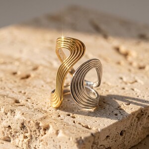 May include: A two-tone ring featuring a gold-colored section and a silver-colored section, both designed with parallel, wavy, grooved lines that create an open, fluid, and asymmetrical structure that wraps around the finger.