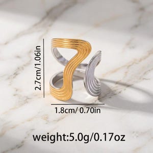 May include: A two-tone ring featuring a gold-colored section and a silver-colored section with wavy, grooved textures. The ring measures 1.06 inches in height, 0.70 inches in width, and has a weight of 0.17 ounces as indicated in the image.